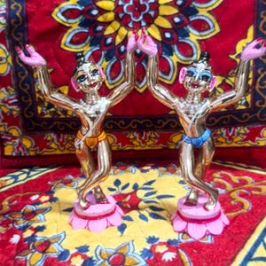 May include: Two gold-colored figurines of dancing figures with raised arms, set on pink lotus flower bases. The figures have painted faces and wear colorful clothing. The background is a red and yellow patterned fabric.