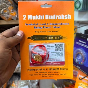 May include: An orange rectangular product card labeled 2 Mukhi Rudraksh, featuring a brown seed bead encased in a clear plastic blister pack with a red string. The card includes text about Lord Ardhnareeshwara, the Moon, and a QR code.