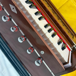 May include: A brown wooden harmonium musical instrument featuring a keyboard with white and black keys, a series of silver-toned metal knobs with red circular bases, and a decorative metal plate with a keyhole on the side of the wooden frame.