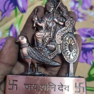 May include: A copper-coloured religious figurine showing a deity in a chariot, with a bird. The base has text in a script. The figurine is held in a hand against a floral background.