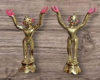 May include: Two gold-colored statues of figures with arms raised, hands painted pink. The figures have detailed facial features and are standing on ornate bases. The background is a wooden surface.