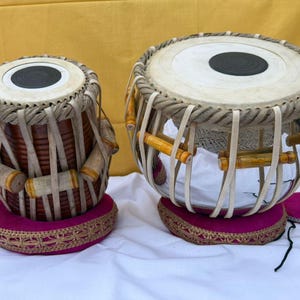 Puede incluir: Un par de tambores tabla que consta de un tambor de madera marrón oscuro más pequeño y un tambor metálico más grande. Ambos tienen parches blancos con centros negros, tensados por correas de cuero claras y clavijas de madera sobre bases magenta.