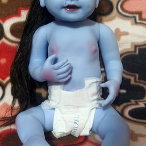 May include: A blue-skinned baby doll with black hair and a white diaper. The doll has a red mark on its forehead and is sitting on a patterned blanket. The doll's features include dark eyelashes and a small mole.