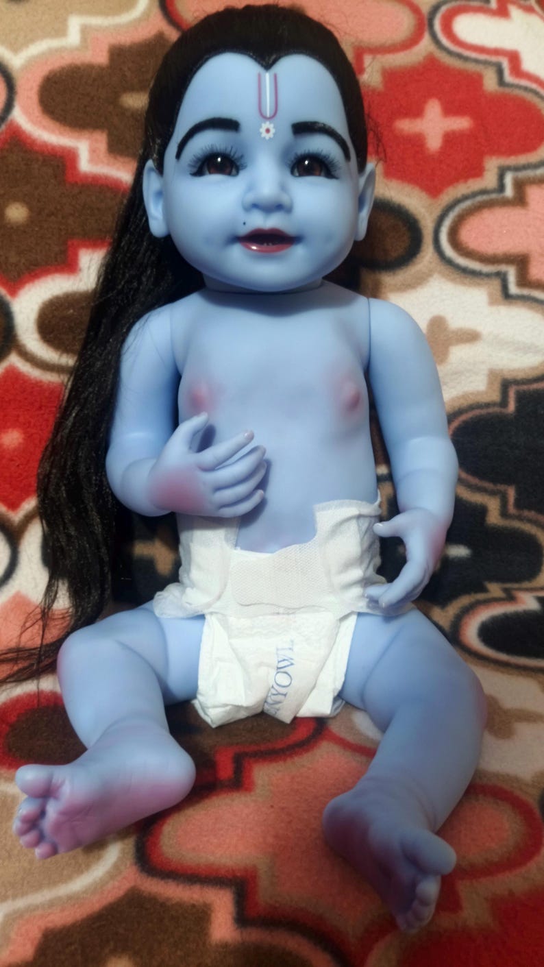 May include: A blue-skinned baby doll with black hair and a white diaper. The doll has a red mark on its forehead and is sitting on a patterned blanket. The doll's features include dark eyelashes and a small mole.