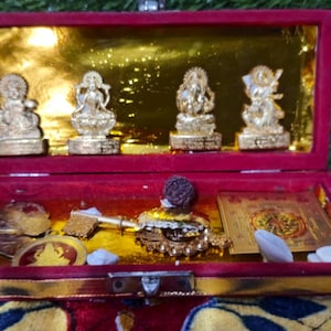 Könnte beinhalten: Eine rote Samtbox mit goldenem Innenraum, die vier kleine Goldstatuen, Münzen und andere religiöse Gegenstände enthält. Die Box ist geöffnet und zeigt den Inhalt vor einem grün-rot gemusterten Hintergrund.