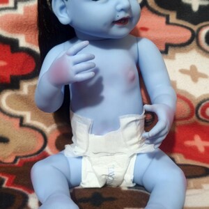 May include: A blue-skinned baby doll with black hair and a white diaper. The doll has a small dot on its forehead and is sitting on a patterned blanket. The doll's skin is a light blue color.