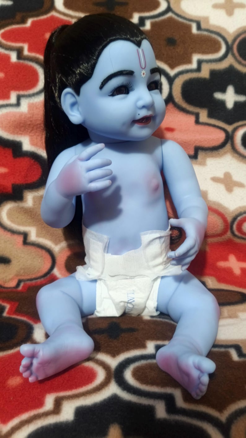 May include: A blue-skinned baby doll with black hair and a white diaper. The doll has a small dot on its forehead and is sitting on a patterned blanket. The doll's skin is a light blue color.