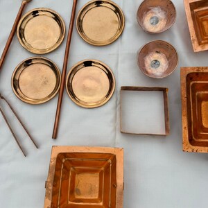 May include: An assortment of copper kitchenware, including small plates, bowls, square molds, and long-handled spoons. The items are arranged on a white surface, showcasing their warm, metallic tones. The collection suggests a focus on culinary or decorative use.