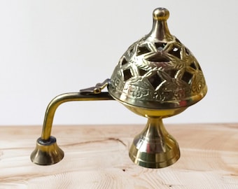 May include: A brass incense burner with a star-patterned lid and a curved handle. The burner has a pedestal base and a small, round finial on top. The metal has a polished, golden finish, and the burner is set against a light wooden surface.