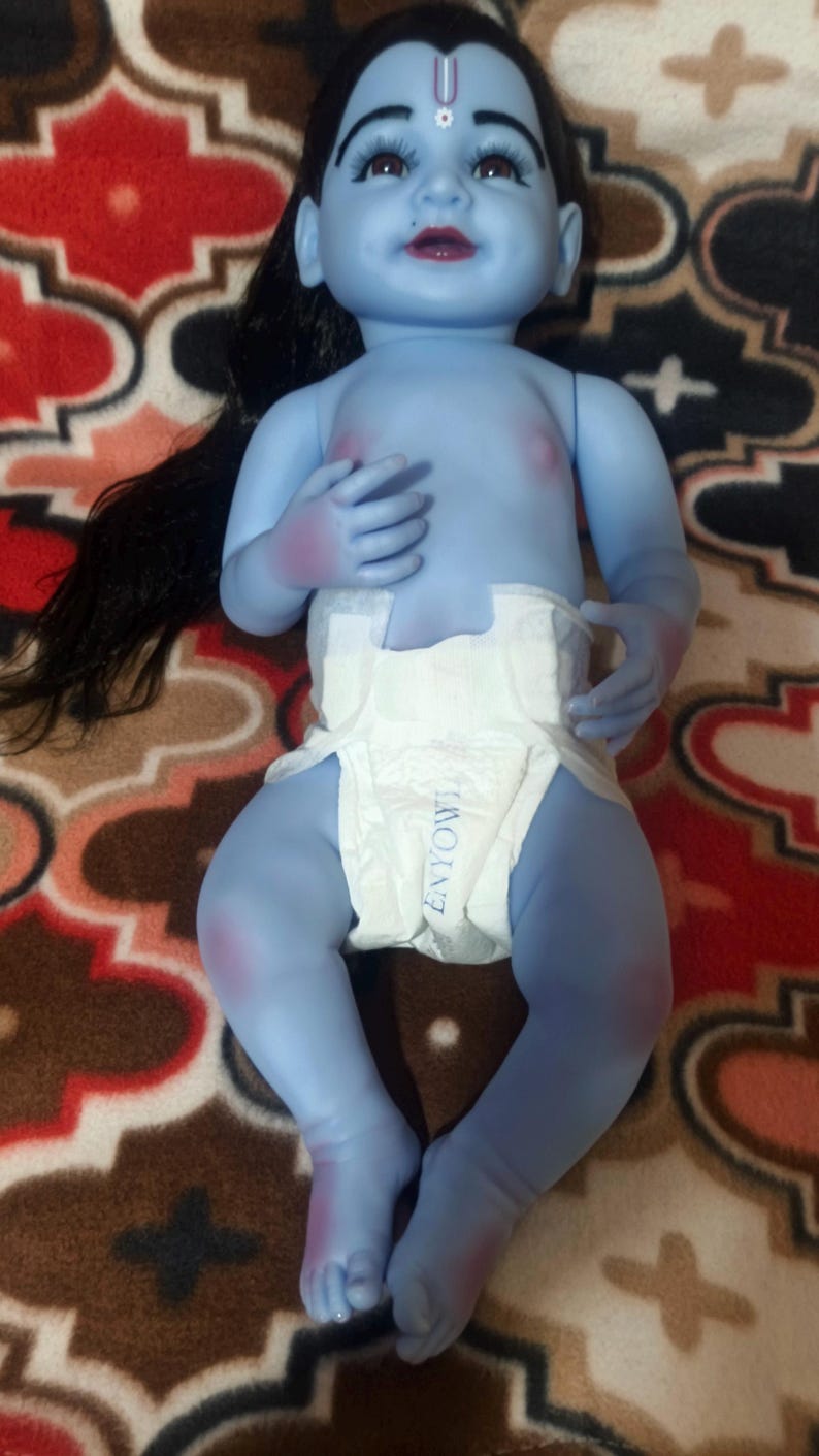 May include: A blue-skinned baby doll with black hair and a white diaper. The doll has a red bindi on its forehead and is lying on a patterned blanket. The doll's eyes are open, and it has a slight smile.