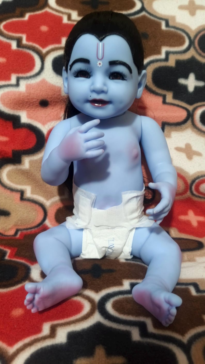 May include: A blue-skinned baby doll with black hair and a white diaper. The doll has a painted face with dark eyes and a red lip color. The doll is sitting on a patterned blanket.
