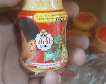 May include: A small, cylindrical container with a red and gold label featuring text in a foreign language and an illustration of a person. The container is held in a person's hand.