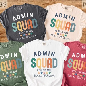 May include: Five t-shirts in various colors: pepper, ivory, moss, white, and crimson. Each shirt features the text "ADMIN SQUAD" in a colorful, retro font, with the phrases "WE GET IT DONE" and "Mrs. Wilson" below. The shirts are likely for administrative staff.