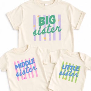 May include: Three cream-colored garments including two t-shirts and one bodysuit. The top shirt features "BIG sister" in green over purple stripes. The bottom left shirt says "MIDDLE sister" in blue over pink stripes. The bodysuit says "LITTLE sister" in blue over green stripes, all with small yellow or pink stars.