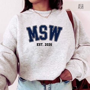 May include: Ash-colored sweatshirt with the letters "MSW" in navy blue block letters, with "EST. 2026" below. The sweatshirt has long sleeves and a crew neck. The model is wearing light blue jeans and a brown belt.