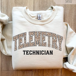 May include: Sand-colored sweatshirt with the words "TELEMETRY TECHNICIAN" in a leopard print design. The sweatshirt is a neutral color and has a crew neck. The text is in a bold, collegiate-style font.