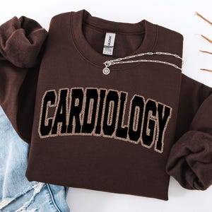May include: A brown sweatshirt with the word "CARDIOLOGY" in large, black, block letters with a leopard print fill. A silver necklace with a small pendant is draped across the sweatshirt. The sweatshirt is folded, with a pair of blue jeans visible.