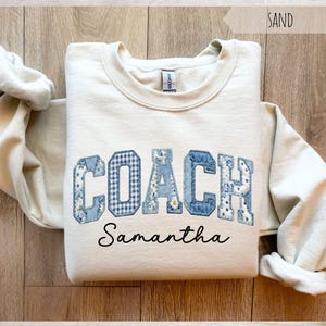 May include: A sand-colored sweatshirt with the word "COACH" in blue patchwork letters. The letter "O" has a gingham pattern, and the other letters have floral patterns. The name "Samantha" is printed below the word "COACH".