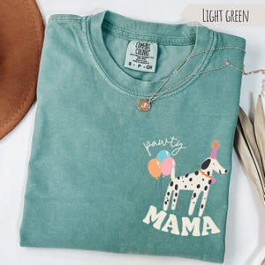 May include: Light green Comfort Colors t-shirt with a graphic of a Dalmatian wearing a party hat and holding balloons. The text "pawty mama" is below the dog. A silver necklace with a pendant is on the shirt.