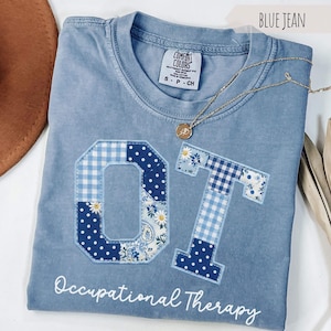 May include: A blue jean colored t-shirt with a Comfort Colors label, featuring the letters OT in a patchwork design of blue and white gingham, polka dots, and floral patterns. The text Occupational Therapy is printed in white below the letters.