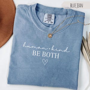 May include: A blue jean Comfort Colors t-shirt with the words "human.kind BE BOTH" in white script and a heart symbol. A gold necklace with a pendant rests on the shirt. The shirt is folded.