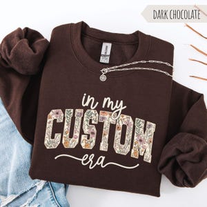 Puede incluir: Sudadera de color chocolate oscuro con el texto "in my CUSTOM era" en un dise&ntilde;o floral. Un collar plateado con un colgante de sol descansa sobre la sudadera. La sudadera est&aacute; doblada sobre una superficie blanca.