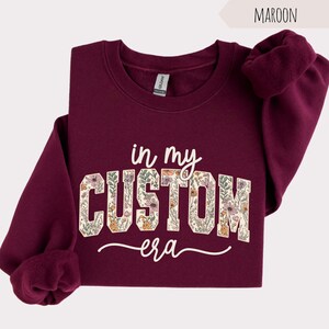 Puede incluir: Sudadera granate con el texto "in my CUSTOM era" en un dise&ntilde;o floral. La sudadera tiene cuello redondo y mangas largas. La palabra "Maroon" est&aacute; en la esquina superior derecha.