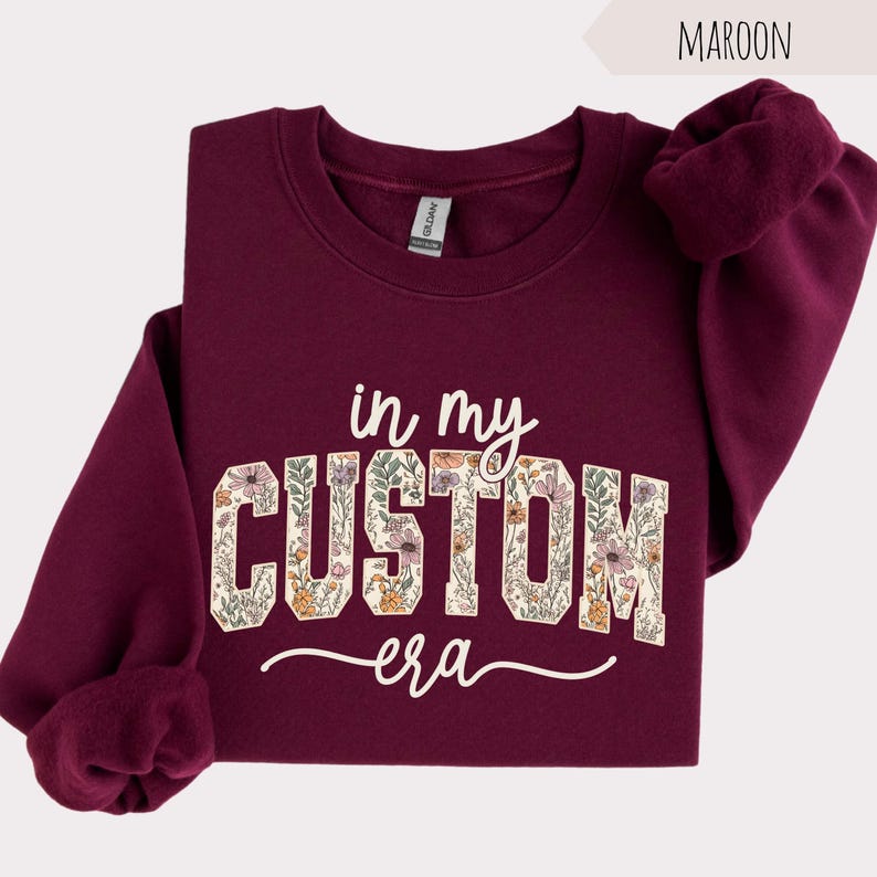 Puede incluir: Sudadera granate con el texto "in my CUSTOM era" en un dise&ntilde;o floral. La sudadera tiene cuello redondo y mangas largas. La palabra "Maroon" est&aacute; en la esquina superior derecha.