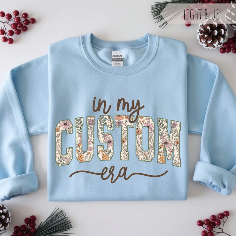 Puede incluir: Sudadera azul claro con la frase "in my custom era" en letras marrones y florales. La sudadera es de cuello redondo y tiene mangas largas. La etiqueta de la marca Gildan es visible.