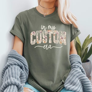 Puede incluir: Camiseta verde oliva con el texto "in my CUSTOM era" en un dise&ntilde;o floral. La camiseta tiene mangas cortas y un corte relajado. El texto est&aacute; en una fuente serif con un patr&oacute;n floral.