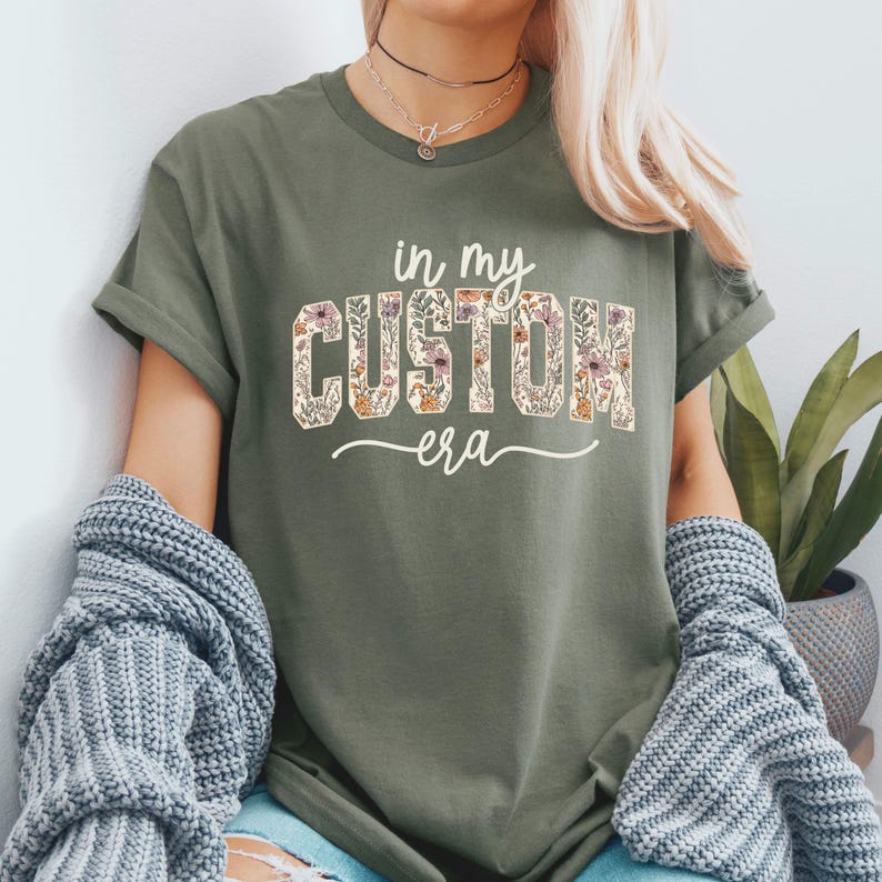 Puede incluir: Camiseta verde oliva con el texto "in my CUSTOM era" en un dise&ntilde;o floral. La camiseta tiene mangas cortas y un corte relajado. El texto est&aacute; en una fuente serif con un patr&oacute;n floral.