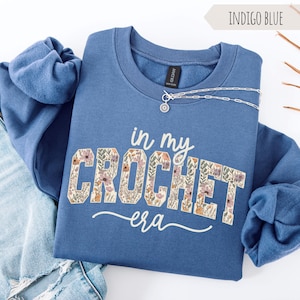 Puede incluir: Sudadera azul &iacute;ndigo con el texto "in my crochet era" en un dise&ntilde;o floral. La sudadera tiene cuello redondo y mangas largas. Un collar plateado con un colgante est&aacute; drapeado en la parte delantera.