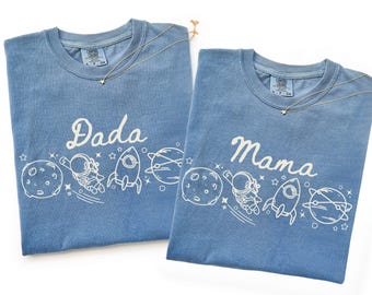 May include: Two blue jean-colored t-shirts with white graphics and text. One shirt says "Dada" and the other says "Mama". Each shirt features a space-themed design with planets, astronauts, and rockets. A gold necklace with a star pendant is on each shirt.