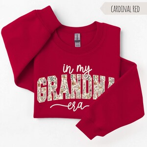 Puede incluir: Sudadera roja cardenal con el texto "in my GRANDMA era" en escritura blanca y estampado floral. La sudadera es de cuello redondo con mangas largas. El texto "Cardinal Red" est&aacute; en una pancarta en la esquina superior derecha.