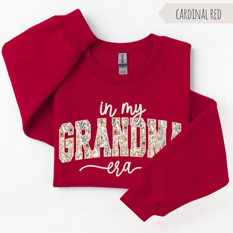 Puede incluir: Sudadera roja cardenal con el texto "in my GRANDMA era" en escritura blanca y estampado floral. La sudadera es de cuello redondo con mangas largas. El texto "Cardinal Red" est&aacute; en una pancarta en la esquina superior derecha.