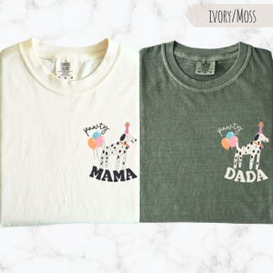 May include: Two t-shirts, one ivory and one moss green, each featuring a Dalmatian dog illustration with a party hat and balloons. The ivory shirt says "Mama" and the green shirt says "Dada" below the dog.