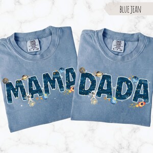 May include: Two blue jean-colored t-shirts with the words "MAMA" and "DADA" in large, space-themed lettering. The letters are filled with a starry night design, and decorated with planets, rockets, and astronauts. The shirts are labeled "Comfort Colors".