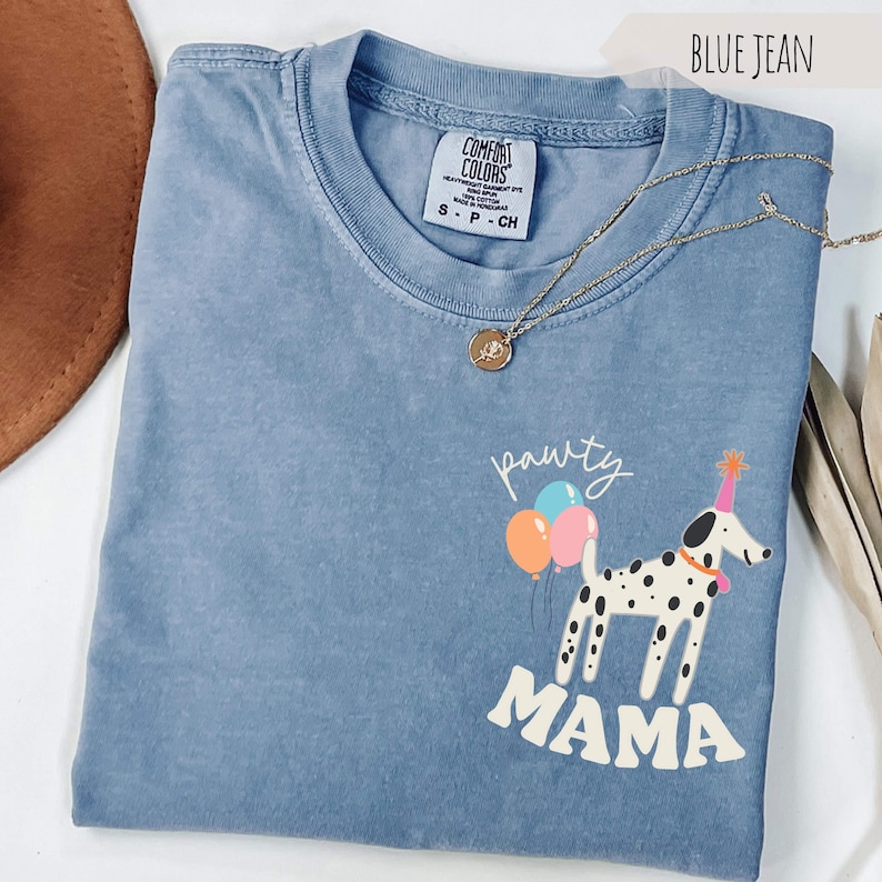 May include: Blue jean colored t-shirt with a graphic of a Dalmatian dog wearing a party hat and holding balloons. The text "pawty MAMA" is printed below the dog. A gold necklace is draped across the shirt.