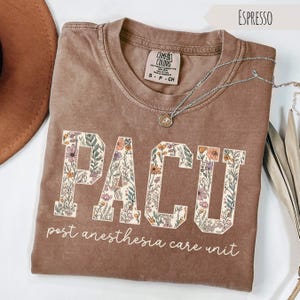 May include: Espresso-colored t-shirt with the letters "PACU" in a floral design. The words "post anesthesia care unit" are printed below the letters. A silver necklace with a pendant is draped across the shirt.
