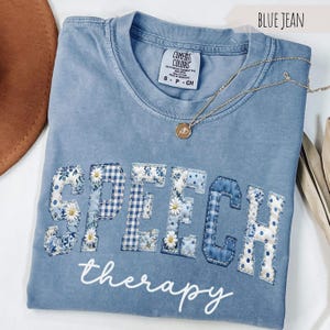 May include: Blue jean-colored Comfort Colors t-shirt with the words "SPEECH therapy" appliqued on the front. The letters are a patchwork of blue floral, gingham, and polka dot fabrics. A gold necklace with a pendant is draped across the shirt.