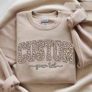 May include: A sand-colored crewneck sweatshirt featuring the word CUSTOM in a varsity-style font with a brown and black leopard print pattern. Below the word CUSTOM, the phrase your text is printed in a black cursive script font.