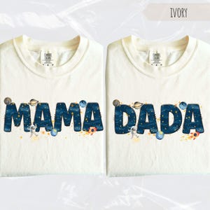 May include: Two ivory-colored t-shirts with the words "MAMA" and "DADA" in a dark blue, space-themed font. Each letter is filled with a starry night design, and the shirts feature illustrations of planets, astronauts, and rockets.