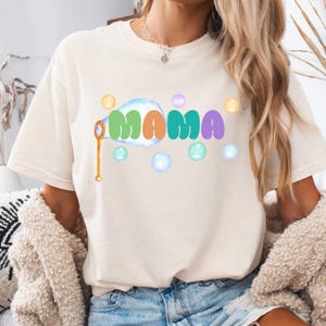 May include: A cream-colored short-sleeve t-shirt featuring the word MAMA in colorful, rounded letters. The design includes a bubble wand and several floating bubbles in shades of blue, green, orange, purple, and yellow surrounding the text.