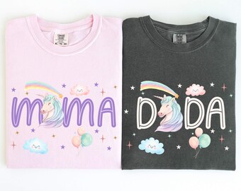 May include: Two t-shirts, one pale pink and one charcoal grey, each with a whimsical design. The pink shirt says "MAMA" in purple script, and the grey shirt says "DADA" in white block letters. Both shirts feature a unicorn, rainbow, clouds, stars, and balloons.