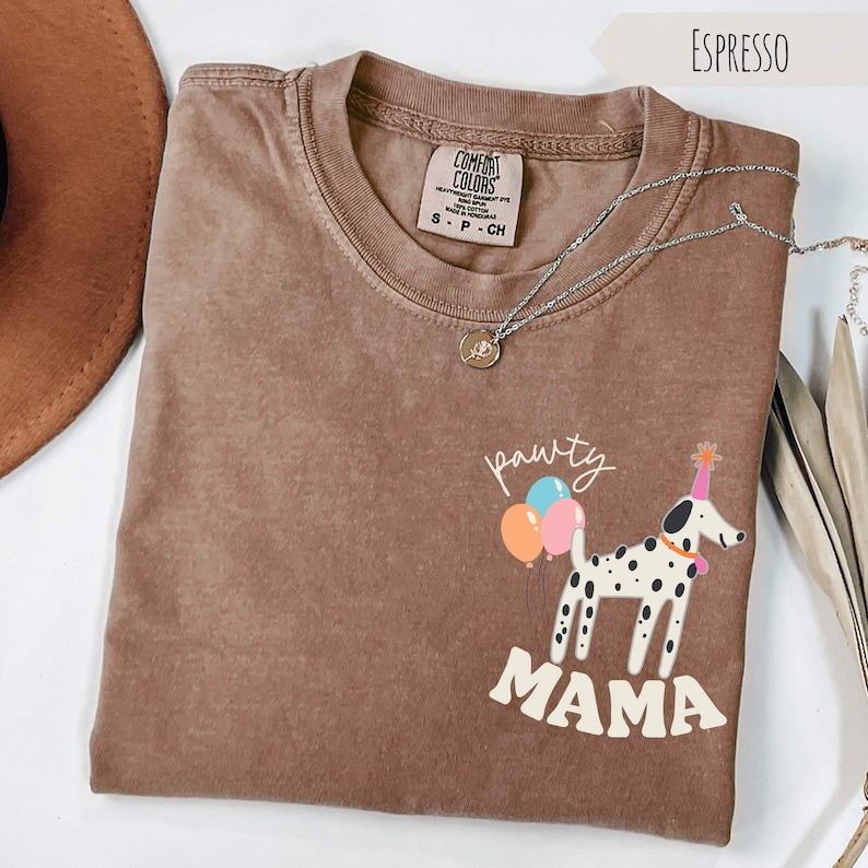May include: Espresso-colored t-shirt with a graphic of a Dalmatian wearing a party hat and holding balloons. The text "pawty mama" is below the dog. A silver necklace with a pendant is on the shirt.