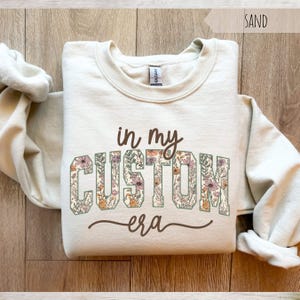 Puede incluir: Sudadera color arena con el texto "in my CUSTOM era" en un dise&ntilde;o floral. La sudadera tiene cuello redondo y mangas largas. La palabra "SAND" est&aacute; impresa en la esquina superior derecha.