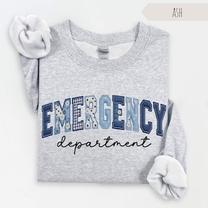 May include: Ash-colored sweatshirt with the words "EMERGENCY department" in a navy blue, patchwork-style font. The letters are decorated with floral and gingham patterns. The sweatshirt has a crew neck and long sleeves.