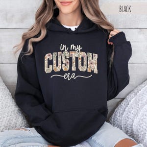 Puede incluir: Sudadera con capucha negra con el texto "in my CUSTOM era" en un dise&ntilde;o floral. La palabra "CUSTOM" est&aacute; rellena con un patr&oacute;n de flores y hojas en tonos beige, naranja y verde. La palabra "era" est&aacute; escrita en cursiva.