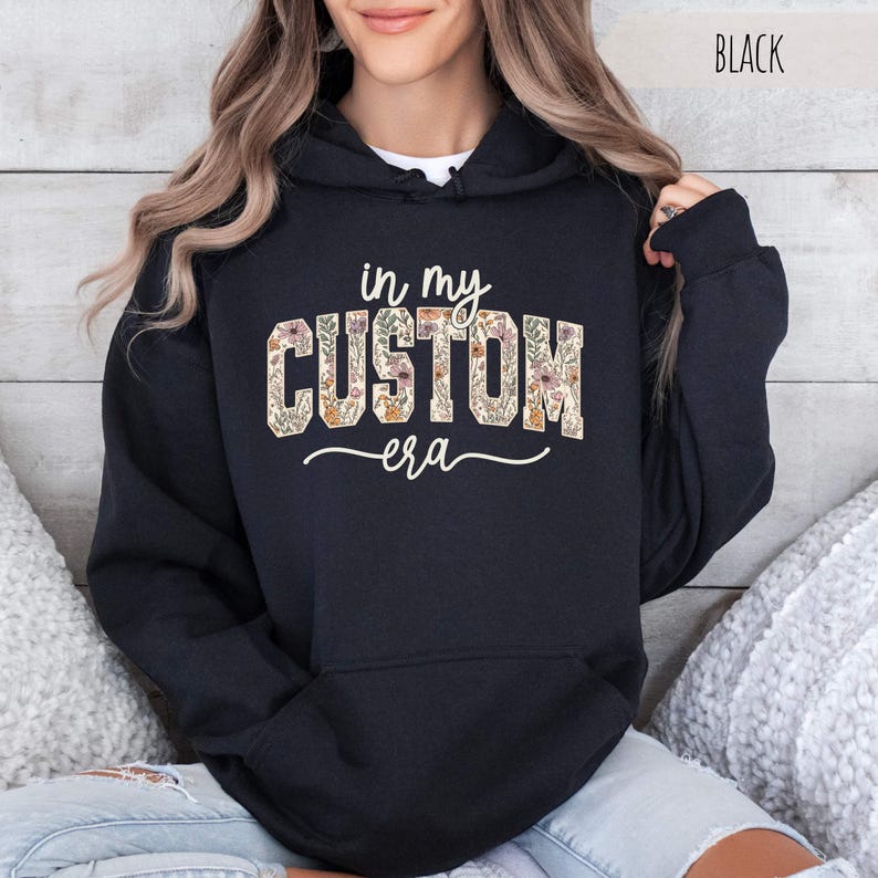 Puede incluir: Sudadera con capucha negra con el texto "in my CUSTOM era" en un dise&ntilde;o floral. La palabra "CUSTOM" est&aacute; rellena con un patr&oacute;n de flores y hojas en tonos beige, naranja y verde. La palabra "era" est&aacute; escrita en cursiva.
