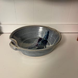 Könnte beinhalten: Eine handgefertigte, blaue Keramikschale mit einer einzigartigen, organischen Form. Die Schale hat eine strukturierte Oberfläche und ein dekoratives, abstraktes Design in Blau-, Lila- und Weißtönen. Sie hat einen kleinen Griff an einer Seite.